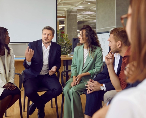 Group of multiethnic people sitting in circle in office interior and listening to mentor sharing his experience, values and leadership mindset.